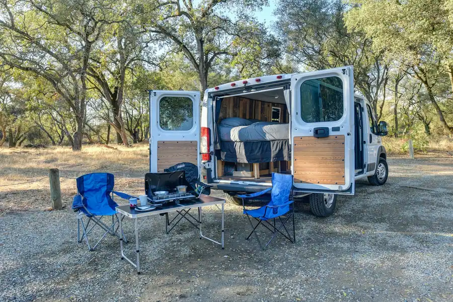 Camper con las puertas trasera y lateral abiertas, sillas plegables y una mesa montada bajo los árboles