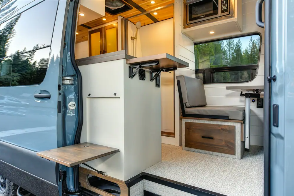 Interior de una camper con banco, mesa plegable y muebles de madera junto a la puerta lateral abierta
