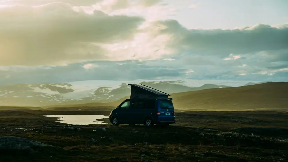 Pequeña camper con techo elevable aparcada en una meseta abierta al atardecer, con colinas nevadas al fondo