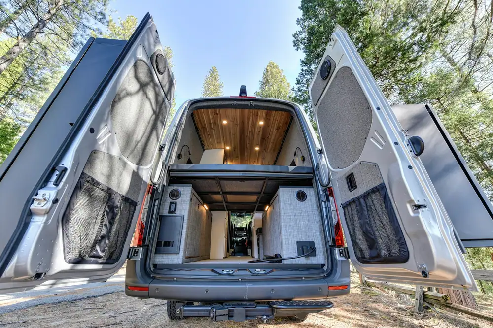 Camper con las dos puertas traseras abiertas, una cama elevada y una zona de almacenaje ordenada debajo