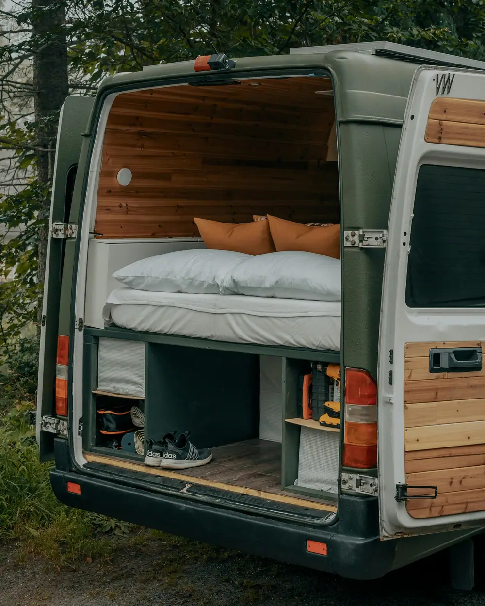 Puertas traseras abiertas de una camper con la cama arriba y un garaje con calzado y bolsas debajo