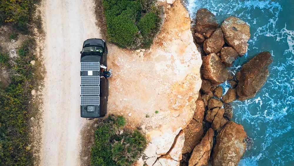 Vista aérea de una camper oscura con placas solares en el techo, aparcada en un camino junto a la costa